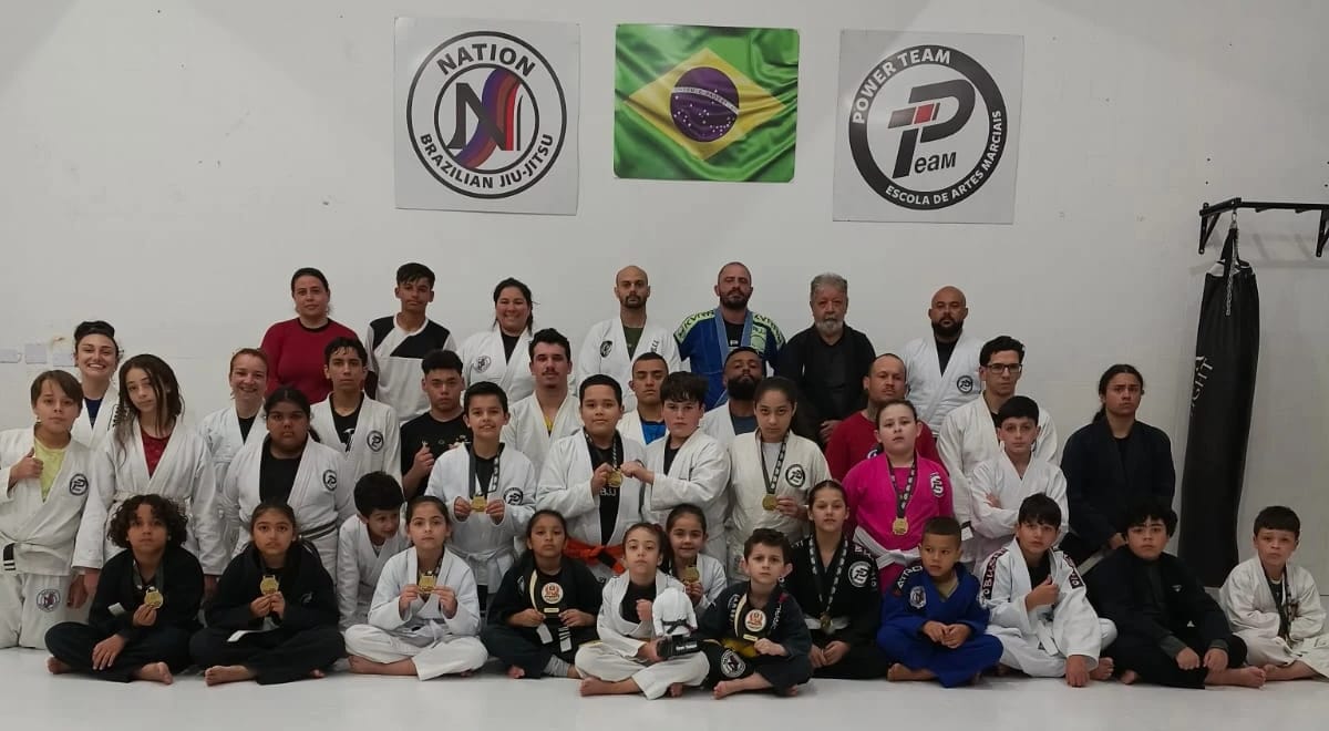 A turma da Nation BJJ, crianças e adultos de kimono, com as medalhas na mão, sob as logos da Nation e da Power Team